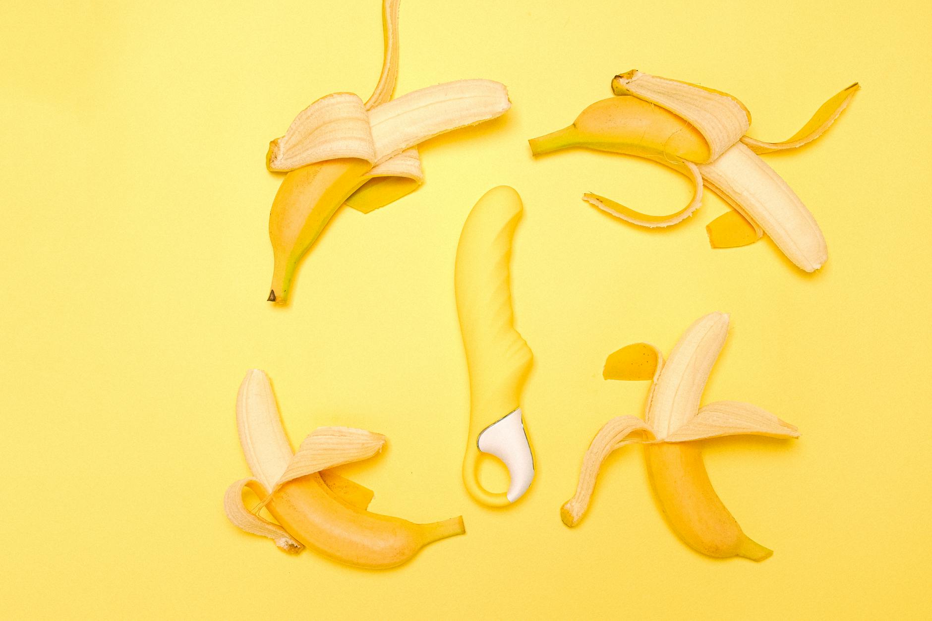 A close-up of a hand holding a yellow silicone vibrator surrounded by fresh fruit on a bright background.